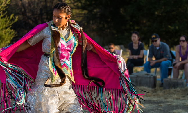 Woman SAMO dancer in traditional clothing performing with audience in the background.