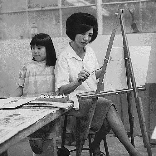 [Detail of] Loretta Graves seated at an easel working on a painting while her daughter stands beside her and looks at other pieces of art on a nearby table.